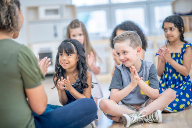 A group of young children sit on the floor in a classroom, clapping and smiling while facing an adult.