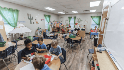 Children sit and work at tables in a classroom decorated with plants and wall art, while others play or read in different activity areas around the room.